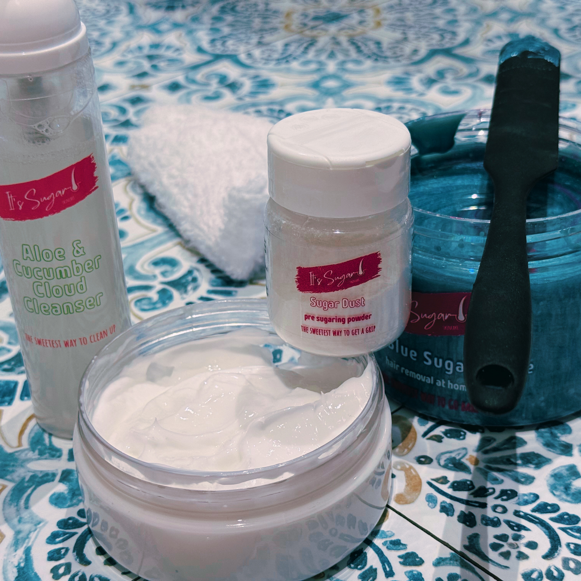 Four products on blue mosaic work surface, Cleanser, Sugar Dust, Moisturiser (lid off) Blue sugar paste for hair removal (lid off) with black applicator on top