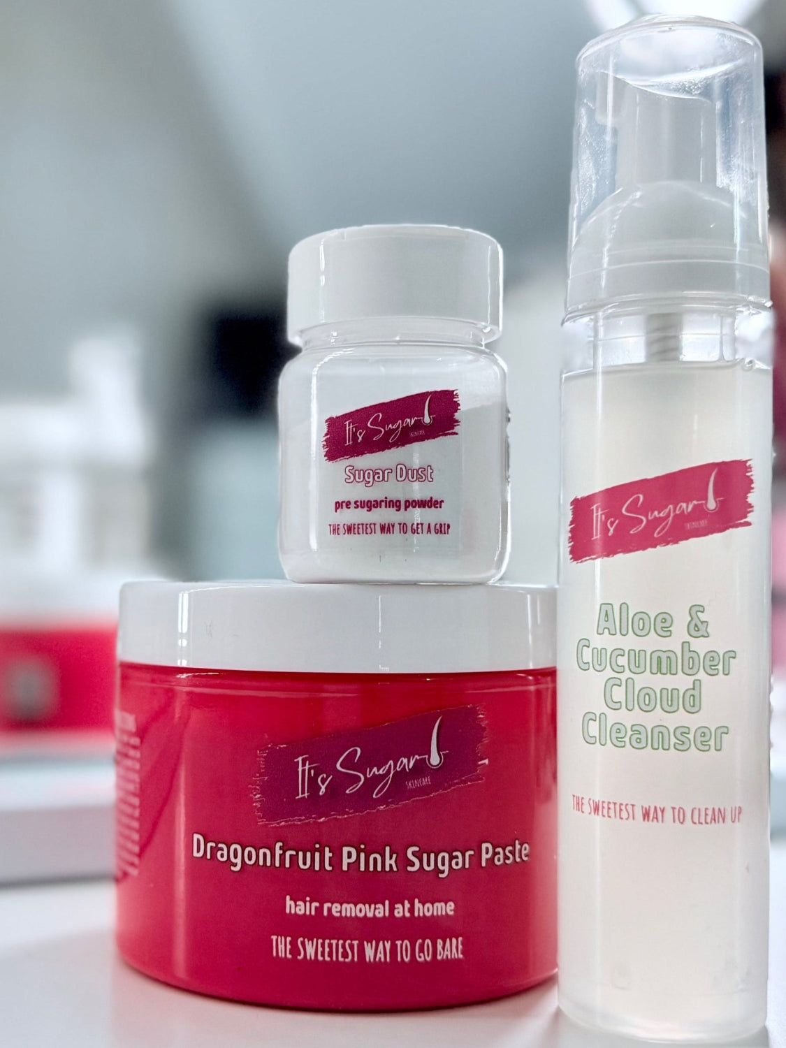 Collection of It's Sugar! hair removal products including a jar, bottle, and container on a white surface in front of a mirror.